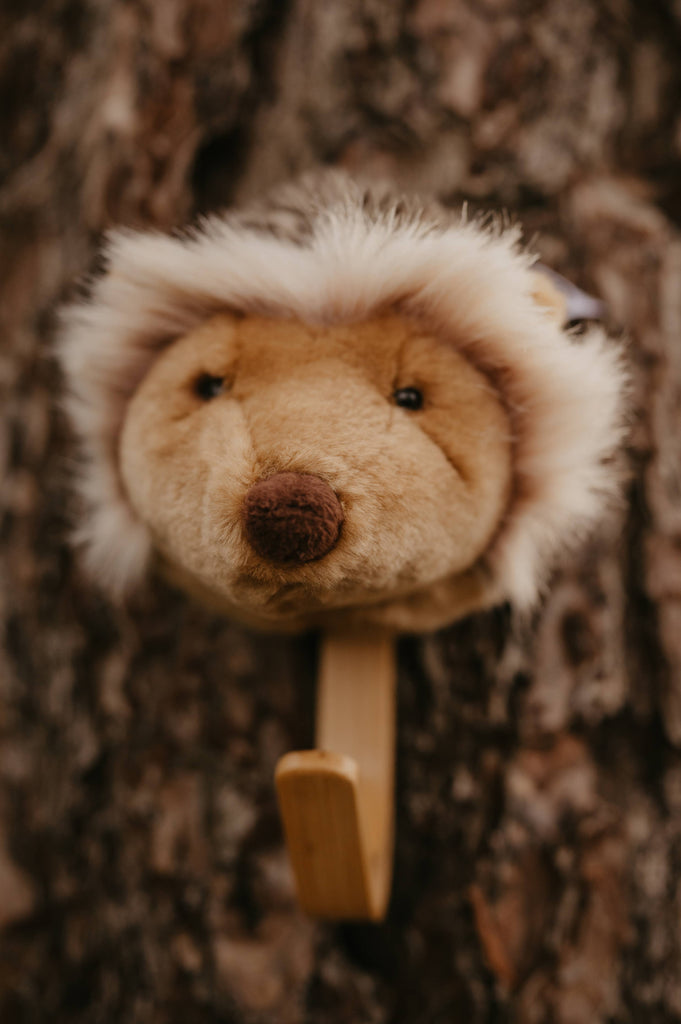 The Coat Hanger - Hedgehog features a plush animal head with a round brown nose, soft beige fur, and a fluffy white mane, displayed against a tree bark background.
