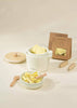 The Wooden Pasta Playset offers a delightful minimalist setup with a white ceramic pot adorned with a wooden lid, holding pasta inside. Complemented by a small bowl of pasta, wooden tongs, and packaged pasta, it perfectly captures the essence of imaginative play against a soft, neutral backdrop.