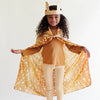 A smiling young child with curly hair stands with arms outstretched in a superhero pose, wearing a brown crown, tan outfit, and Sarah's Silks Animal Print Playsilk - Fawn as a cape against a plain background.