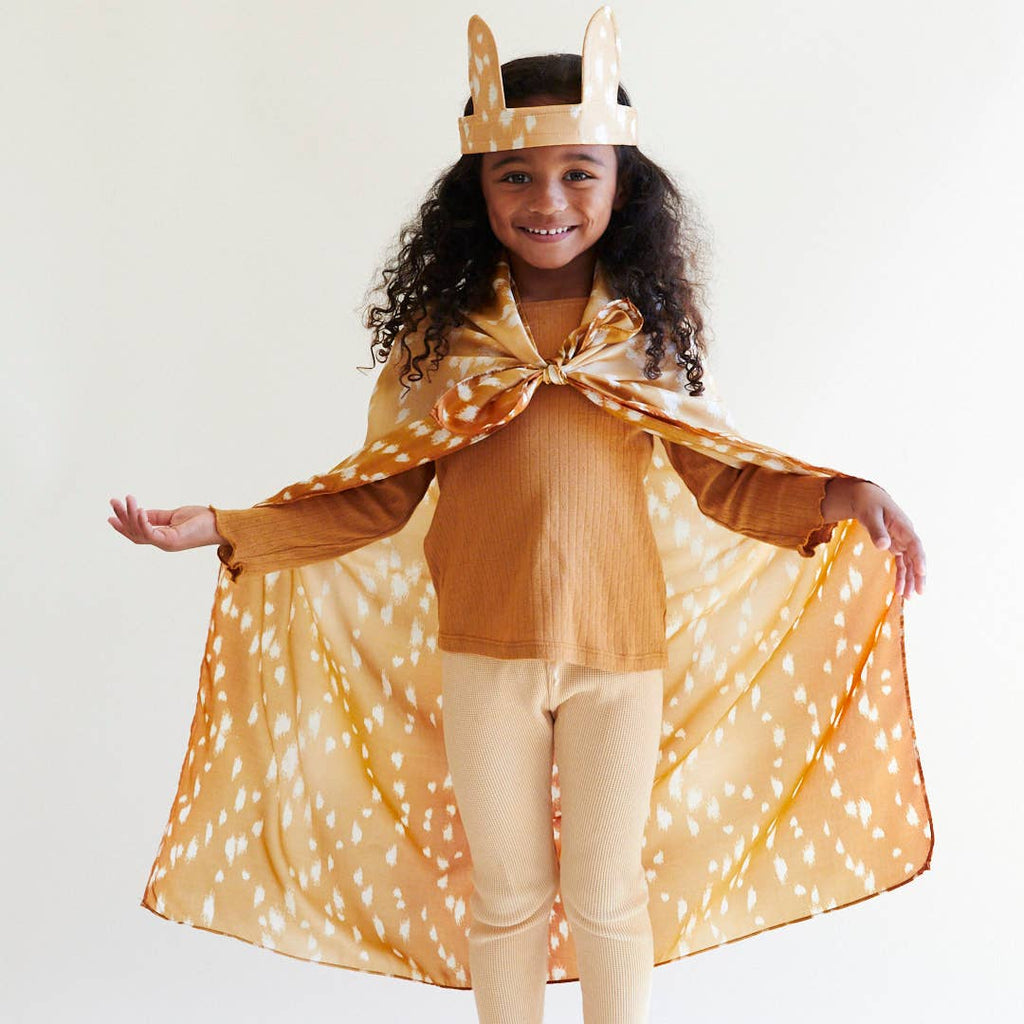 A smiling young child with curly hair stands with arms outstretched in a superhero pose, wearing a brown crown, tan outfit, and Sarah's Silks Animal Print Playsilk - Fawn as a cape against a plain background.