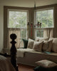 A Coral & Tusk Christmas Cheer Wreath Pillow among a collection of embroidered linen holiday pillows arranged on a window seat under a candelabra chandelier.
