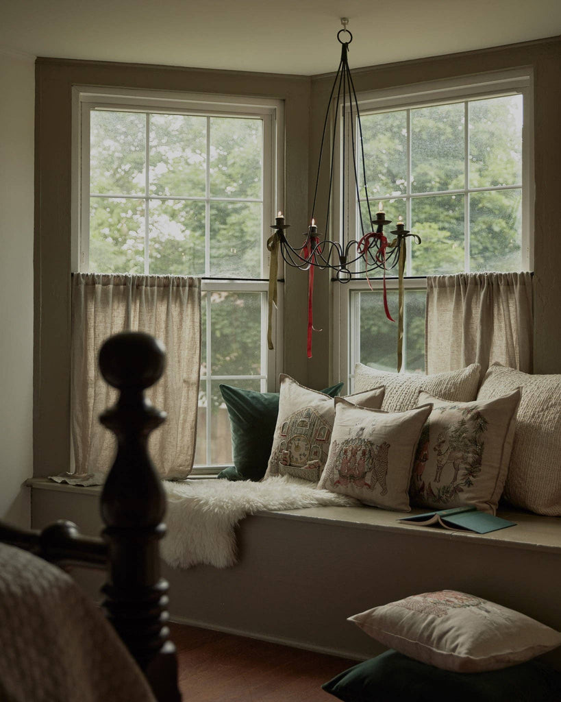 A Coral & Tusk Christmas Cheer Wreath Pillow among a collection of embroidered linen holiday pillows arranged on a window seat under a candelabra chandelier.