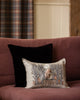 A maroon sofa with a black cushion and a Coral & Tusk Spooky Forest Pocket Pillow featuring a scarecrow, pumpkins, and trees, in a cozy room with plaid curtains and wood-paneled walls.
