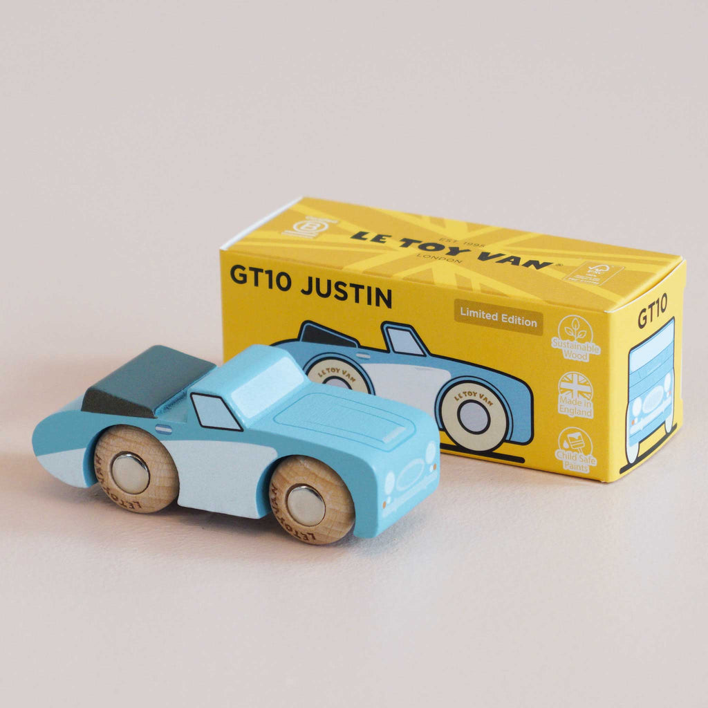 A small blue wooden car rests in front of a yellow box labeled “No. 1 GT10 Justin | Made in England” and “LE TOY VAN.” The box displays an illustration of the classic car along with several icons, making it a delightful keepsake for any collection.