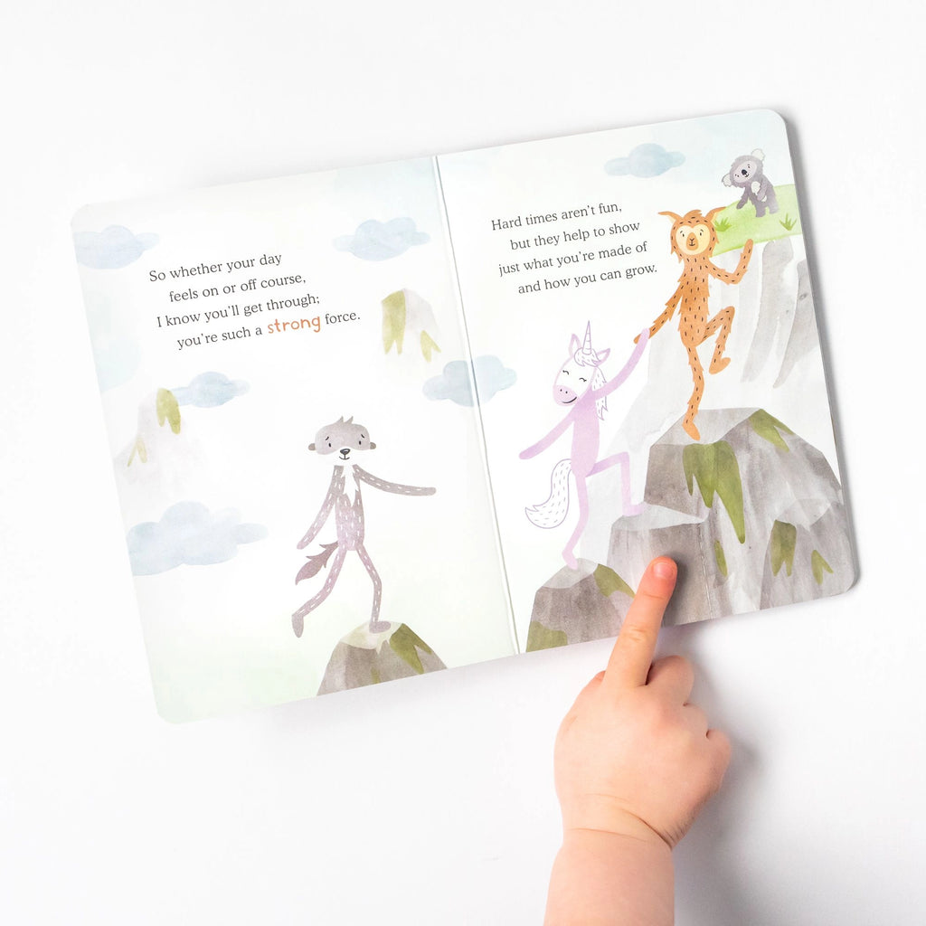 A child's hand points to an open Slumberkins Alpaca Kin + Lesson Book On Stress Relief, showing whimsical animals on a mountain, with comforting text about strength, growth, and anxiety management on the pages.
