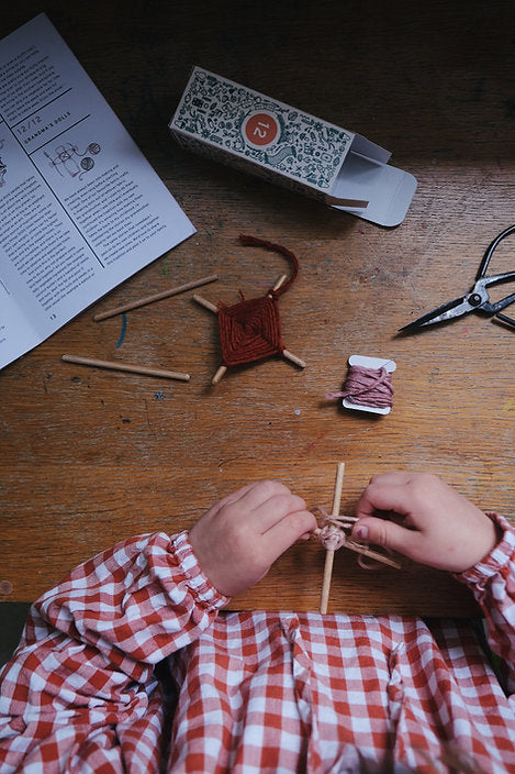 A child in a plaid shirt knits with yarn and needles, surrounded by a Grapat Advent Calendar (Final Sale) and scissors on a wooden table.