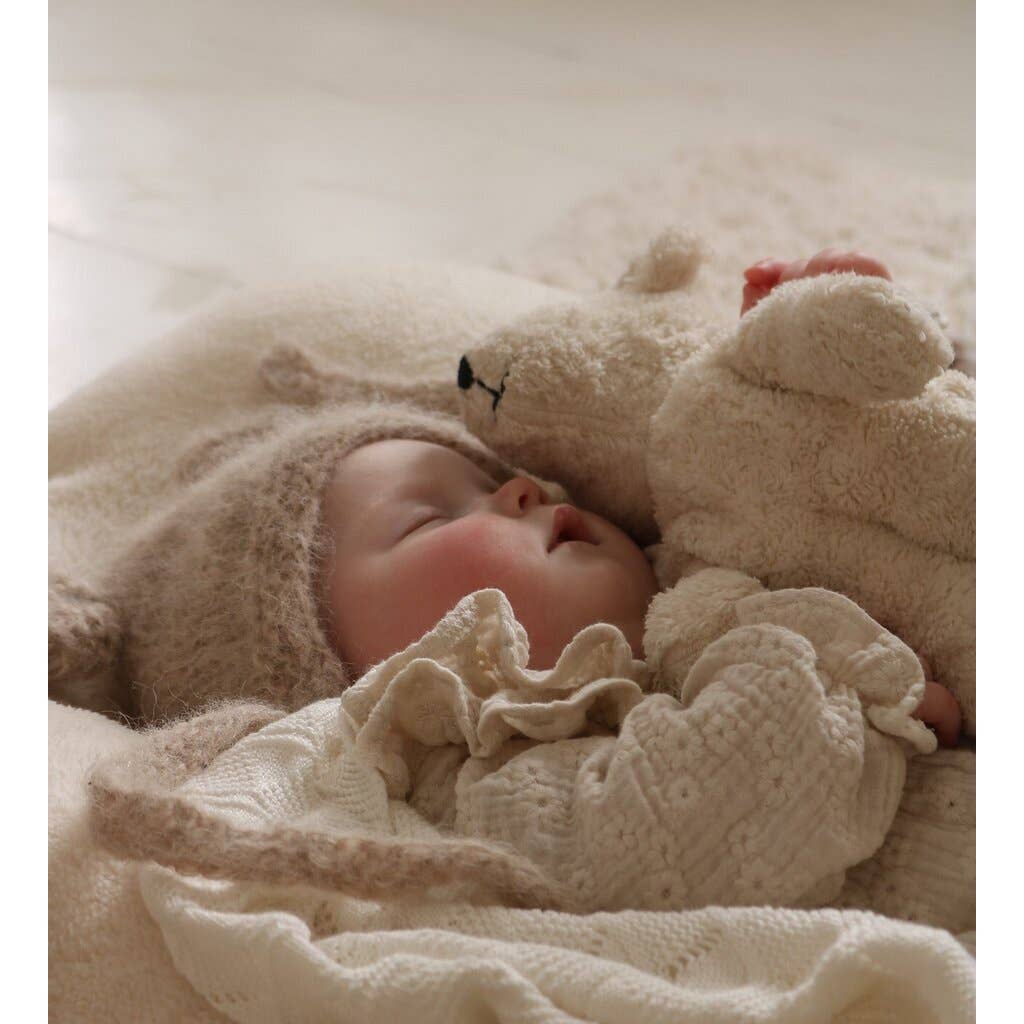 A baby in a cozy knitted beige outfit and hat sleeps peacefully, cuddling the Senger Naturwelt Stuffed Animal - White Bear on a cream-colored blanket.