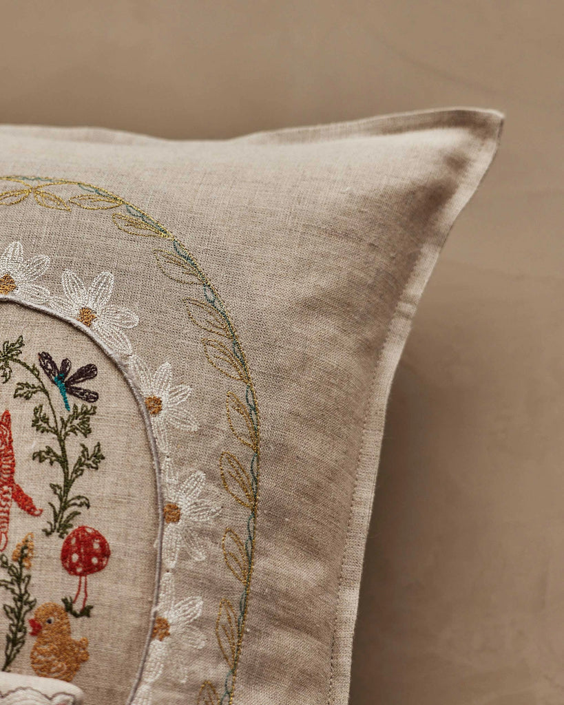 A close-up of the Coral & Tusk Sugar Egg Pocket Pillow in beige linen, showcasing detailed embroidery of white flowers, a red mushroom, and green leaves—an ideal neutral accent for Easter décor.
