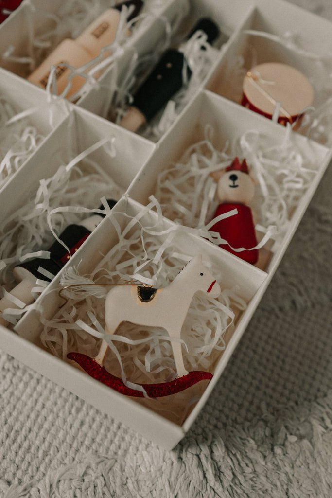 The Sabo Concept Christmas Ornaments set includes festive, hand-painted wooden figures—a white rocking horse, drum, nutcracker, and a red-clad figure—in a compartmentalized white box filled with white shredded paper.