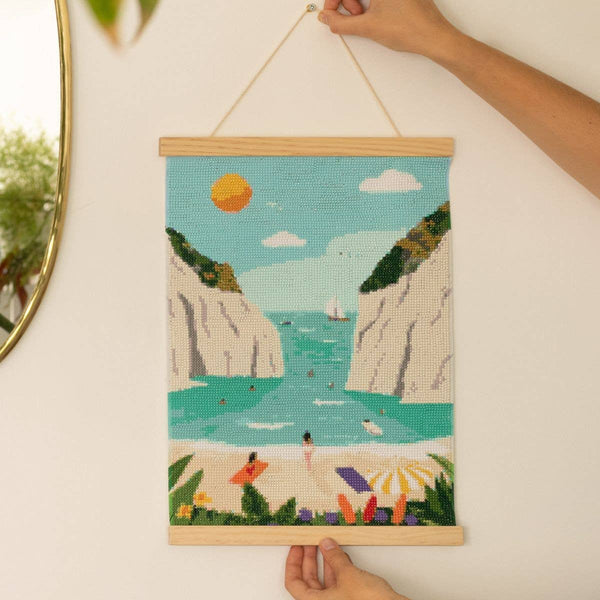 A hand displays the "Diamond Painting Kit - Stiniva Beach by Maja Tomljanovic," showing a vibrant Mediterranean cove with cliffs, sailboats, and people under umbrellas. This seaside artwork hangs on a wall by a string.