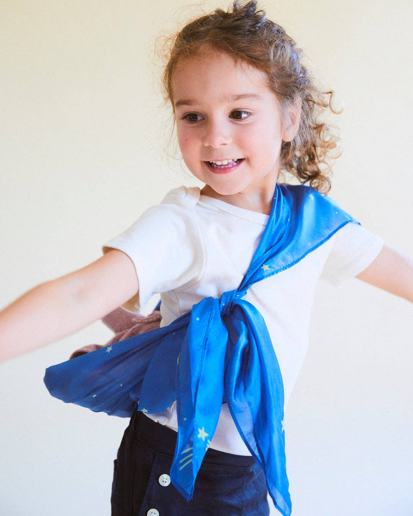 A young child smiles and spins, wearing a white shirt, dark pants with buttons, and Sarah's Silks Enchanted Playsilk - Starry Night tied around their shoulders—perfect for open-ended play. The background is light and plain.