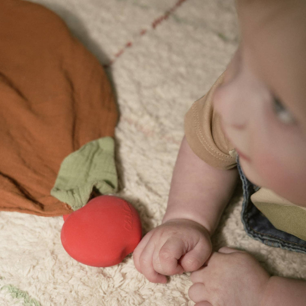 A baby lies on a soft, textured surface, holding a red apple-shaped teether with a green leaf. Nearby is the Pepa the Apple Big Doudou, an orange and green organic cotton comforter. The baby's face is partially visible and out of focus.