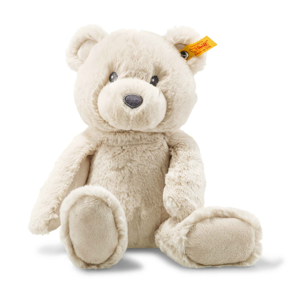 A Steiff, Bearzy Teddy Bear Stuffed Plush For Baby, 11 Inches with soft beige fur, black eyes, and a small black nose sits upright. The bear has a yellow tag with red text attached to its left ear, ensuring it’s gentle enough for a baby's soft skin.