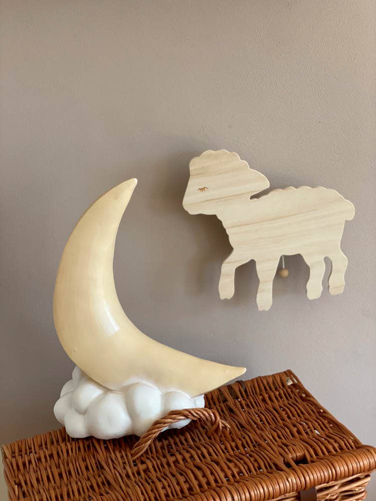 A decorative wooden sheep hangs on a neutral-toned wall above a wicker basket, while a Crescent Moon Lamp, resembling a nursery lamp, rests in front of the basket on white clouds, adding to the whimsical design.