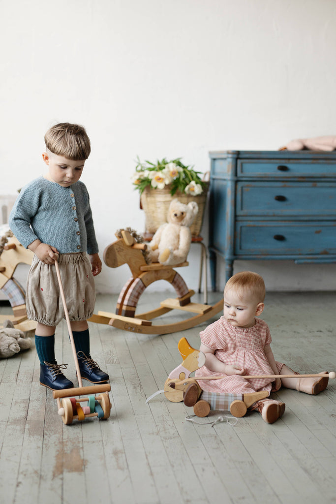 A young boy in a blue sweater and beige shorts pushes the Handmade Wooden Toy, Push Toy Rattle, while a baby in pink sits with a pull-along duck. A teddy bear, rocking horses, and a blue dresser set the scene with natural materials.