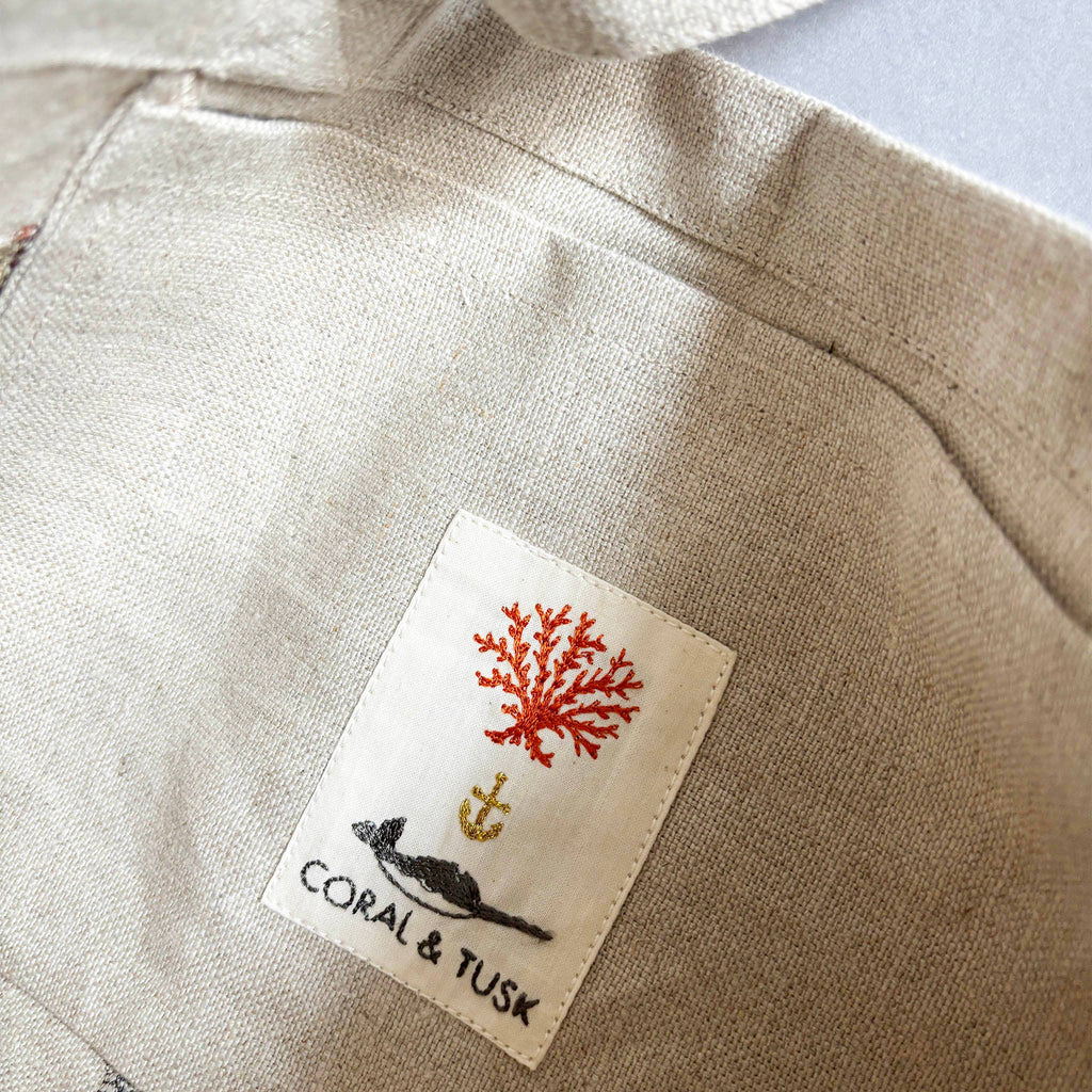 Close-up of a fabric with a sewn label featuring red coral, a small anchor above a black whale, and the words "Coral & Tusk." The textured fabric could perfectly complement the Cats Tote Bag, potentially revealing charming feline faces inside its pocket.