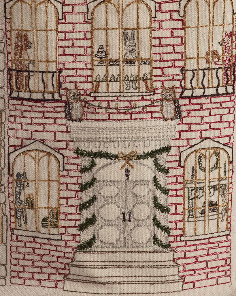 The "Home for the Holidays Bucket" features embroidery of a brick building with arched windows, festive double doors, cats and shelves inside, plus an owl above the entrance for added holiday charm.