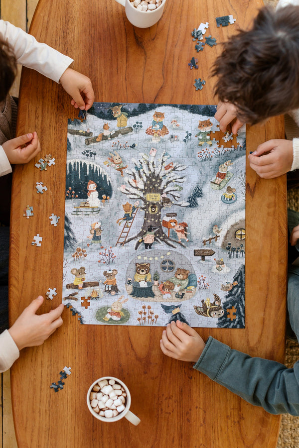 Three children work together to assemble the Midnight Mail 500 Piece Puzzle, featuring whimsical animal characters, on a wooden table as they enjoy mugs filled with marshmallows.