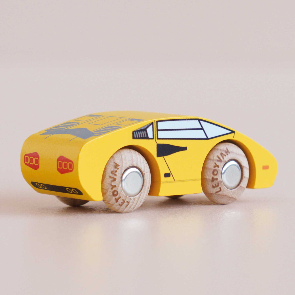 A yellow wooden No. 14 GT80 Lando sports car with black detailing and wooden wheels, featuring the word “LETOVIAN” on the wheels, is displayed on a light beige surface. Handcrafted in England, this toy supercar boasts a sleek design and a child-safe finish.