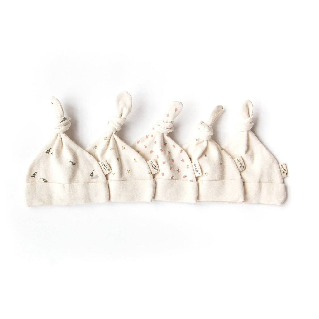 Five cream-colored Organic Baby Hat – Schooner hats with knotted tops are lined up side by side, each with a small tag and delicate floral or sprout patterns—ideal as organic newborn essentials.