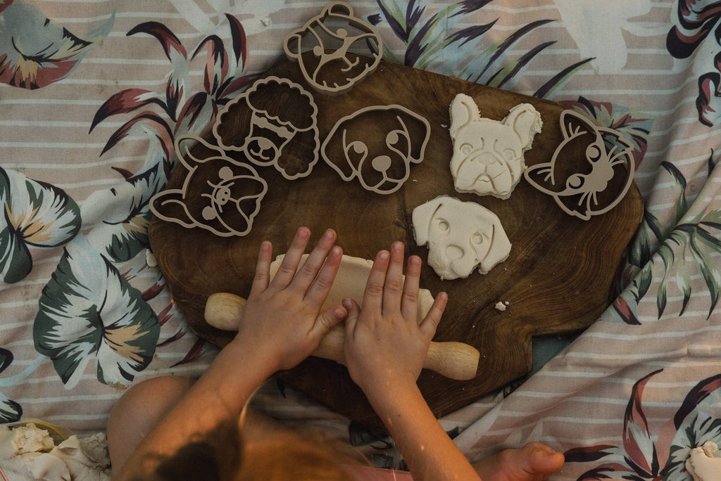 A child uses a rolling pin on dough beside a wooden board, where animal-shaped creations made with the Pet Friends Faces Cutter Set - Eco Playdough Tool are showcased on a floral-patterned surface—inviting sensory play with these engaging playdough tools.