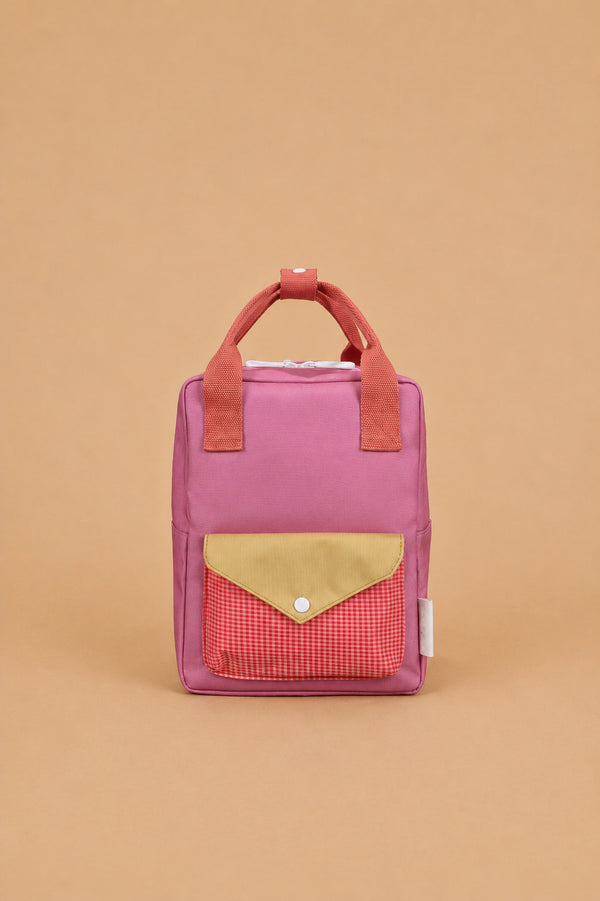 A Sticky Lemon Backpack Small - Envelope in Rosebay Pink is displayed against a plain beige background, showcasing its small rectangular waterproof design with pink material, red handles, a yellow flap, a red checkered front pocket, and adjustable shoulder straps.
