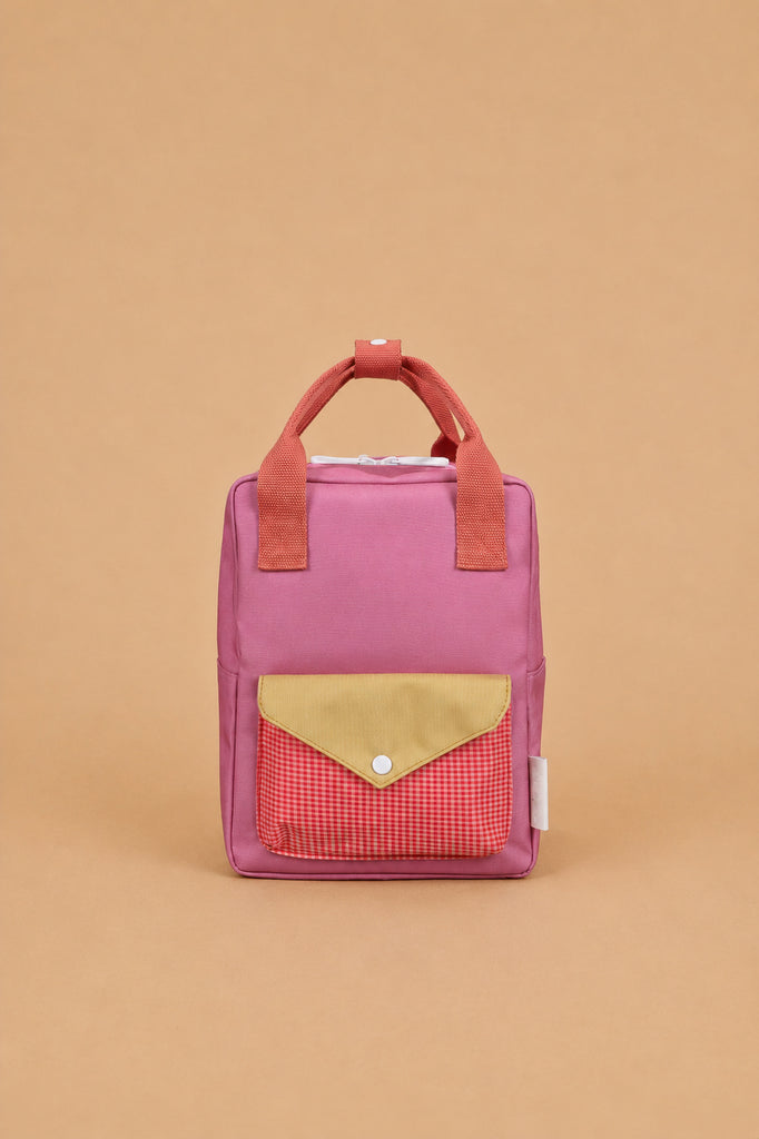 A Sticky Lemon Backpack Small - Envelope in Rosebay Pink is displayed against a plain beige background, showcasing its small rectangular waterproof design with pink material, red handles, a yellow flap, a red checkered front pocket, and adjustable shoulder straps.
