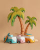 Three VW Beach Bus Pink toy vans, each with a magnetic camper top and a surfboard, are parked before two wooden palm trees on a beige background, creating a playful beach scene.
