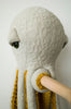 A soft, textured, light gray BigStuffed Octopus - Pop with large green eyes and mustard yellow tentacle underparts is hanging on a wooden rod. The toy, featuring long fluffy tentacles reminiscent of the underwater world, is displayed against a plain white background.