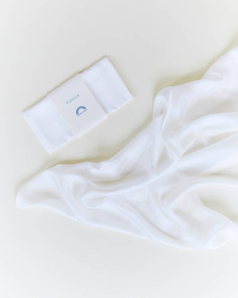 Sarah's Silks Solid Color Playsilk in white is displayed on a light surface beside its "PLYNEL" package featuring a blue half-circle—perfect for open-ended play and creative moments.