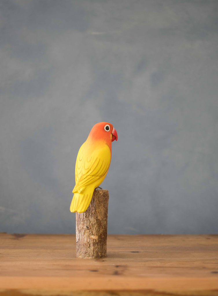 The Hand Carved Ornament - Lovebird - 7.5"H features a vibrant yellow and orange bird figurine perched on sustainable softwood, displayed on a wooden surface with a simple blue-gray background.