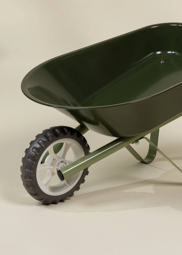 A close-up view of the Heritage Wheelbarrow – Liane, a green children’s wheelbarrow with a single black and white wheel, set against a plain beige background.