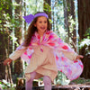 A young girl wearing a purple headband and a vibrant Sarah's Silks Magical Creatures Playsilk - Pink Unicorn smiles joyfully while running through a sunlit forest of tall trees, embracing open-ended play.