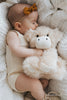 A baby in a cream outfit and brown headband sleeps, hugging Snuggle Bunnies - Hilda The Highland Cow, the perfect plush cuddle companion, on a soft white blanket.