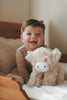 A smiling baby sits on a bed with Snuggle Bunnies - Hilda The Highland Cow, the perfect cuddle companion. Dressed in beige, the baby adds warmth to the cozy room—a delightful choice for a memorable baby shower gift.