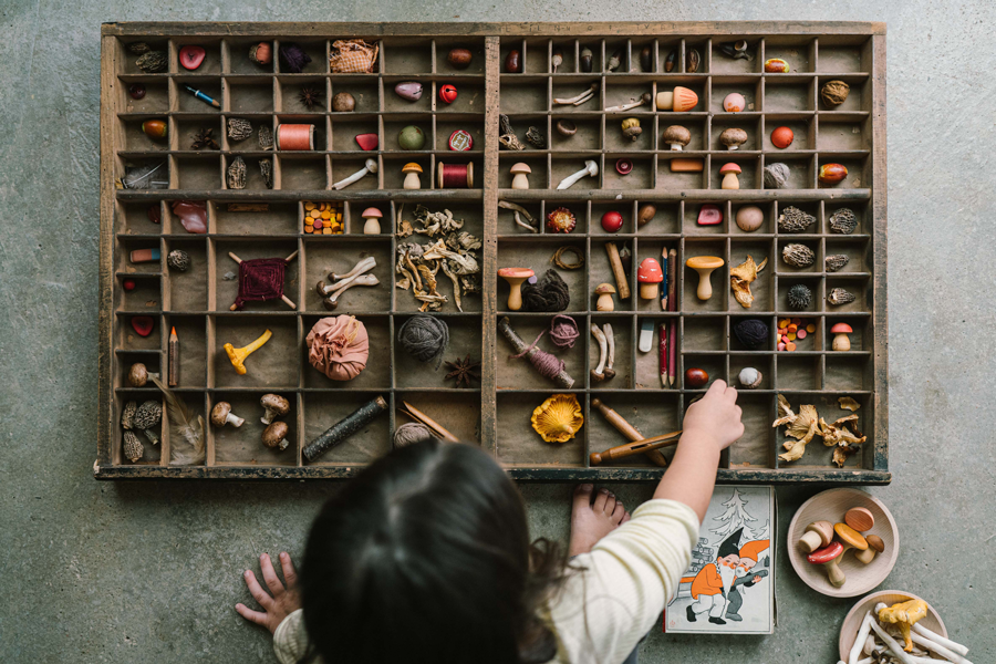 A child plays with the Grapat Play Set - Mushroom Grove, arranging wooden mushrooms and hand-painted toys in a wooden tray. Nearby, there’s a bowl and an animal art card to inspire imaginative play.