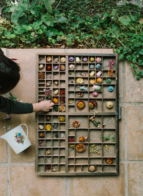 A child arranges the Grapat Play Set - Flower Meadow in a wooden tray outdoors on stone tiles, surrounded by green plants—an inviting scene for imaginative play.
