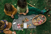 Three children sit on a blanket outdoors, enjoying a picnic with apples, berries, and snacks. A wicker basket holds more fruit and the Grapat Play Set - Fruits, inviting imaginative play on the green grass with scattered apples nearby.
