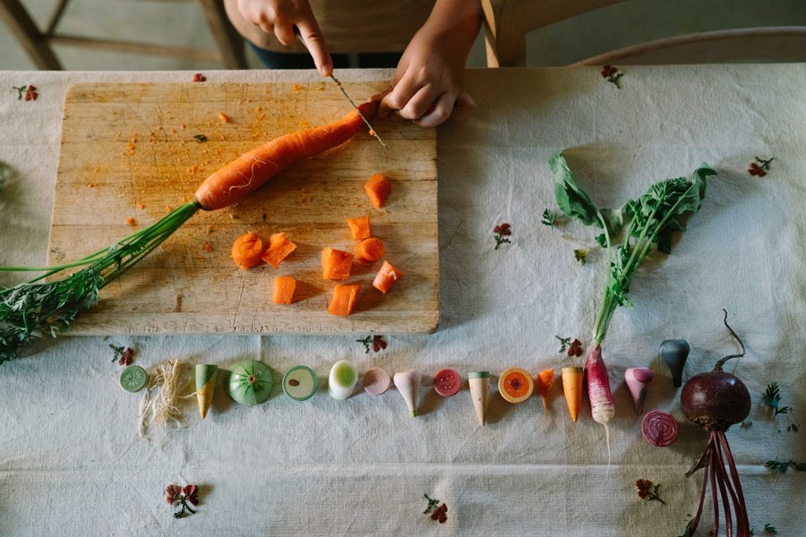 Someone slices a large carrot from the Grapat Play Set - Veggies on a wooden cutting board, with assorted colorful wooden vegetables and their cross-sections neatly displayed on a tablecloth below.