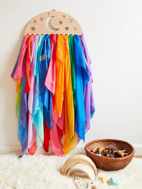 A colorful display of translucent Sarah's Silk Large Star Playsilks in rainbow hues, hanging from a red oak MDF moon and stars motif holder against a white wall, with a wicker basket of toys and a mirror on.