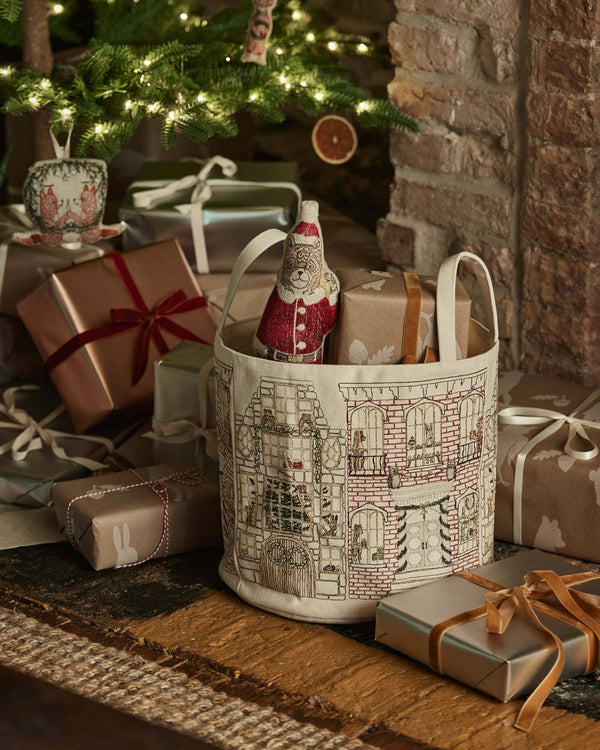 A festive scene with wrapped presents and the Home for the Holidays Bucket—featuring city-style Christmas decor, holding gifts and a Santa doll—is set near a lit Christmas tree by a brick fireplace.