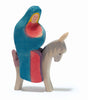 The "Ostheimer Mary on Donkey" wooden figurine, evocative of classic Ostheimer toys, portrays a figure clad in a blue and red cloak, holding a child while riding a gray donkey. Handcrafted from sustainable materials, the figurine is painted with simple, bright colors and features minimalistic design elements that lend it a charming folk art appearance.