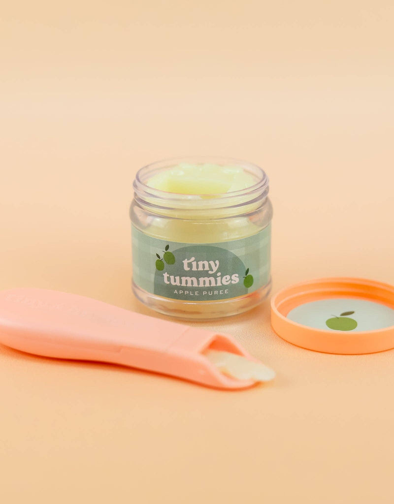 A small jar of Tiny Tummies apple jelly food with the lid removed shows off the light green puree inside. The jar features a green label decorated with apples. Nearby, a peach-colored spoon—reminiscent of a magic spoon for tiny taste buds—holds a sample against a soft peach background, ideal for your little doll eats moment.