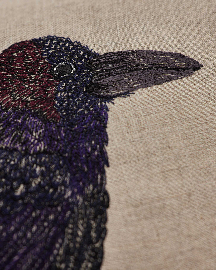 Close-up of the Coral & Tusk Blackbird with Acorn Pillow showcases intricate embroidery of a blackbird on light beige linen, featuring detailed black, purple, and dark red stitching.