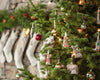 A decorated Christmas tree showcases fabric animal ornaments and assorted colored baubles, with the Santa Bear Ornament dressed in a red suit prominently standing out. In the background, stockings are hung on a stone wall, enhancing the festive mood and hinting at upcoming Christmas gifts.