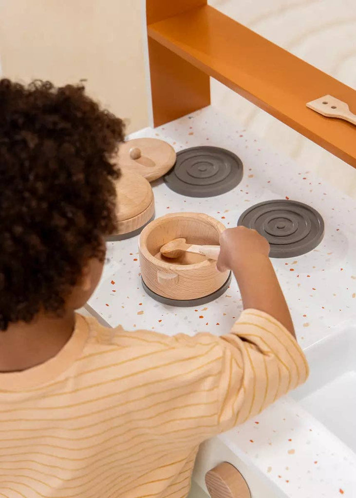 A child with curly hair, wearing a yellow striped shirt, plays with a wooden play kitchen and uses the Wooden Pots and Pans Playset (6 Pcs) on the stove.
