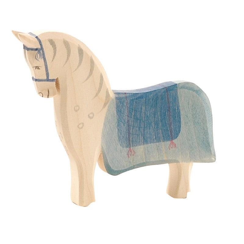 The Ostheimer Horse with Saddle stands with its head slightly lowered. It is painted in a light natural wood color, featuring blue accents such as a blue mane, harness, and a blue blanket over its back. Perfect for imaginative play, this wooden toy horse showcases a simple and rustic design.