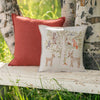 Two decorative pillows rest on a weathered white bench outdoors: one is rust-colored, and the other is a Coral & Tusk Woodland Living Tree Pillow, featuring embroidered forest animals among birch trees and tall green grass.