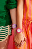 Two people hold hands—one wears green stripes with a Mini Kyomo Watch - Lavender Field in purple, the other wears orange and pink with a pink watch. They’re standing against a pink background.