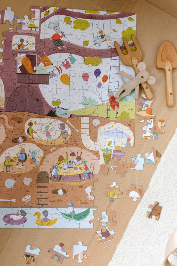 The Beneath the Branches 200 Piece Puzzle with animal characters lies partially assembled on wood beside a plush mouse and two wooden utensils. Matte pieces, some loose, are scattered near the edge of a white rug.